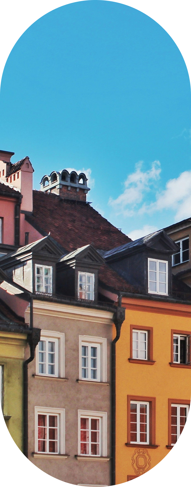 Zbliżenie na fasady i dachy kolorowych, historycznych kamienic, prawdopodobnie z polskiego lub środkowoeuropejskiego starego miasta. Kamienice mają okna ze szprosami, w tym charakterystyczne mansardowe okna wychodzące z ciemnego, spadzistego dachu. Na szczycie dachu widoczna jest metalowa, ozdobna kopuła kominowa. Tło stanowi jaskrawo niebieskie niebo z kilkoma białymi chmurami.